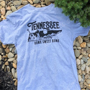 May include: A light blue t-shirt with a black graphic design featuring a guitar, a person, a mountain range, and the text "Tennessee Home Sweet Home".