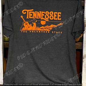 May include: A dark grey t-shirt featuring an orange graphic design of the state of Tennessee. The graphic includes a guitar, a dog, and the text "Tennessee, The Volunteer State".