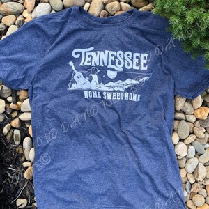 May include: A blue t-shirt with a white graphic design that says "Tennessee Home Sweet Home". The graphic features a dog sitting on a guitar with mountains and a moon in the background.