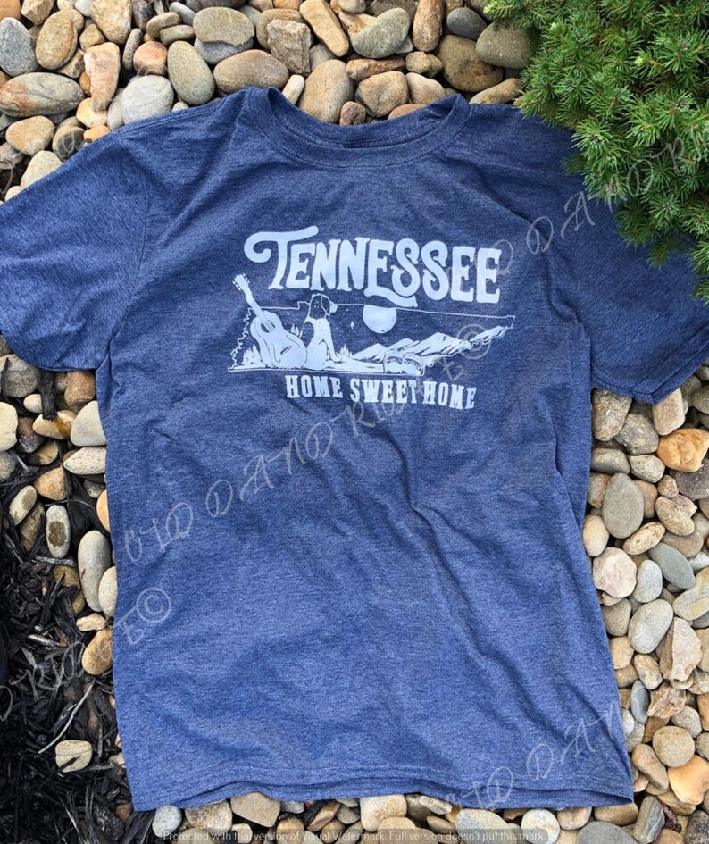 May include: A blue t-shirt with a white graphic design that says "Tennessee Home Sweet Home". The graphic features a dog sitting on a guitar with mountains and a moon in the background.