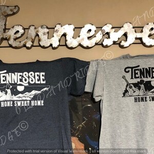 May include: Two t-shirts featuring a graphic design of the state of Tennessee with a guitar and the text "Tennessee Home Sweet Home". One t-shirt is dark grey and the other is light grey.