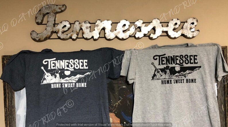 May include: Two t-shirts featuring a graphic design of the state of Tennessee with a guitar and the text "Tennessee Home Sweet Home". One t-shirt is dark grey and the other is light grey.