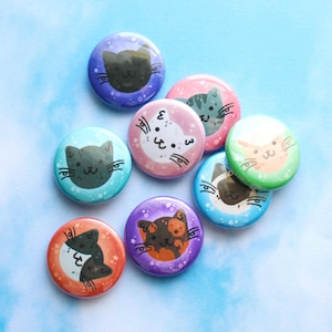 May include: A collection of eight circular pin-back buttons featuring various cat face illustrations. Each button displays a different feline color pattern, including black, white, grey, calico, and brown, set against vibrant backgrounds with small star motifs.