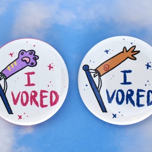 May include: Two white round buttons with blue, pink and orange cartoon arms and hands. The buttons say "I Vored" in pink and blue letters.