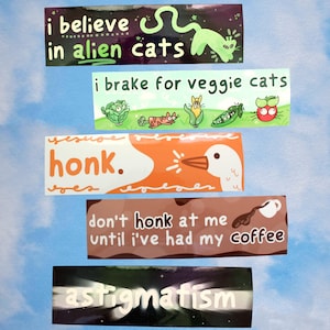 May include: A collection of rectangular stickers with humorous phrases and illustrations. The stickers include text like "I believe in alien cats," "I brake for veggie cats," and "don't honk at me until I've had my coffee." The background is a light blue sky.
