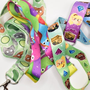 May include: Four colorful lanyards with different cartoon designs. One lanyard features a green background with avocado and cat illustrations. Another lanyard has a pink background with colorful cartoon monsters. A third lanyard has a green background with a treasure chest and gems. The final lanyard has a purple background with cartoon characters and gems.