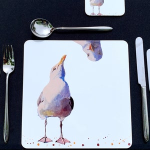 May include: A white placemat featuring a blue and white watercolour illustration of two seagulls. One seagull is standing upright, while the other is upside down.