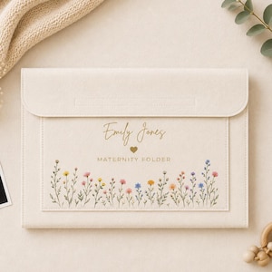 Puede incluir: Una carpeta de maternidad rectangular de color crema con cierre de solapa. La parte frontal presenta el texto Emily Jones, un pequeño icono de corazón y las palabras MATERNITY FOLDER, junto a una colorida ilustración en acuarela de flores.