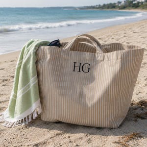 Könnte beinhalten: Eine beigefarbene Tasche mit vertikalen weißen Streifen und den aufgestickten schwarzen Initialen HG auf der Vorderseite. Ein hellgrünes Handtuch mit weißen Streifen und weißem Fransenbesatz liegt über der Seite der Tasche, neben einer schwarzen Sonnenbrille.