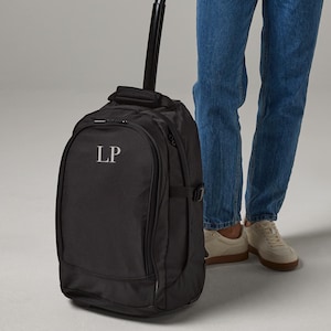 May include: A black rolling backpack with the initials "LP" in white. The backpack has a telescoping handle. The person is wearing a blue button-down shirt, a white vest top, and blue jeans.