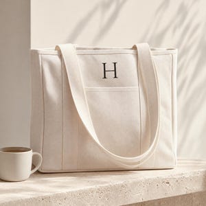 Pode incluir: Uma bolsa tote de lona na cor creme com a letra H bordada em tom escuro no centro da parte frontal. A bolsa possui duas alças longas de ombro e um bolso horizontal na frente. À esquerda, encontra-se uma caneca de cerâmica a combinar.