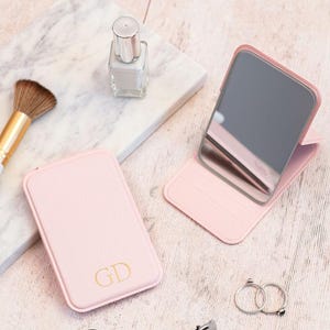 Peut inclure: Deux miroirs de poche roses, dont un ouvert et un fermé, avec des initiales dorées. Un pinceau de maquillage, du vernis à ongles, un recourbe-cils et des bagues sont également présents. Le miroir est sur une surface claire.