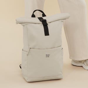 Puede incluir: Una mochila gris claro de rollo superior con correas negras y una hebilla negra. La mochila tiene un bolsillo frontal con las iniciales "BF" impresas en negro.