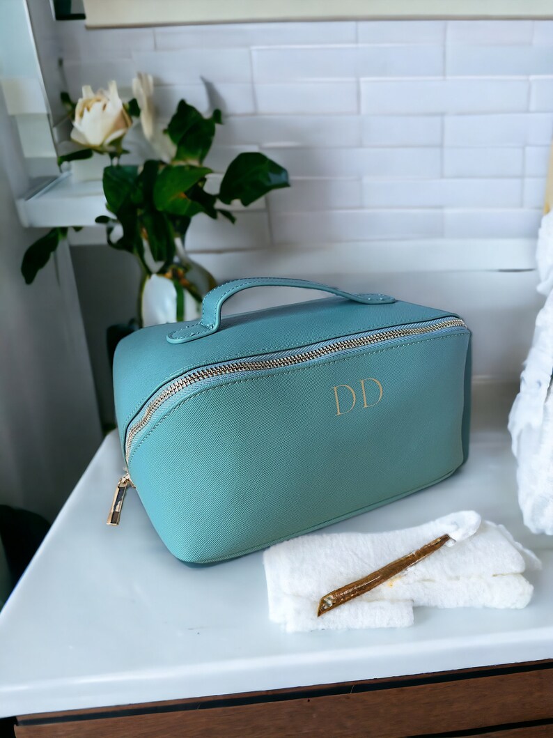 May include: A teal blue zippered travel makeup bag with a gold zipper and handle. The bag has the initials "DD" in gold lettering on the front.