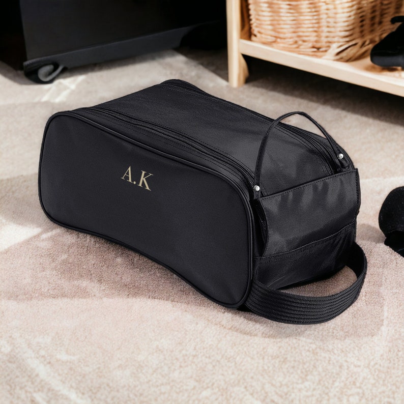 Puede incluir: Bolsa de zapatos negra con cierre de cremallera y asa. La bolsa tiene un peque&ntilde;o bolsillo lateral y las iniciales "A.K." bordadas en la parte delantera.