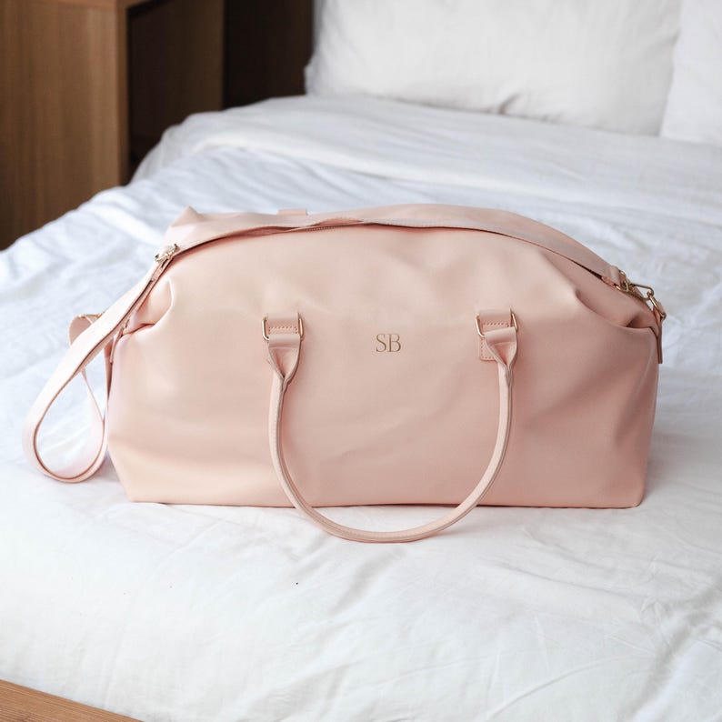 Puede incluir: Una bolsa de viaje de cuero rosa p&aacute;lido con dos asas superiores y una correa de hombro larga. La bolsa tiene una cremallera dorada y las iniciales "SB" est&aacute;n grabadas en la parte delantera.