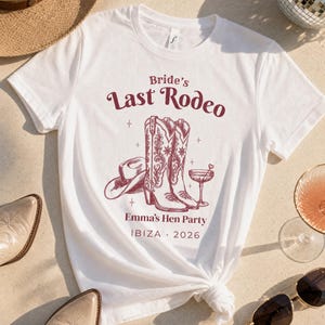 Può includere: Una t-shirt bianca a maniche corte con una grafica bordeaux raffigurante un cappello da cowboy, un paio di stivali western e un bicchiere da cocktail. Il testo recita "Bride's Last Rodeo", "Emma's Hen Party" e "IBIZA 2026" in carattere bordeaux.