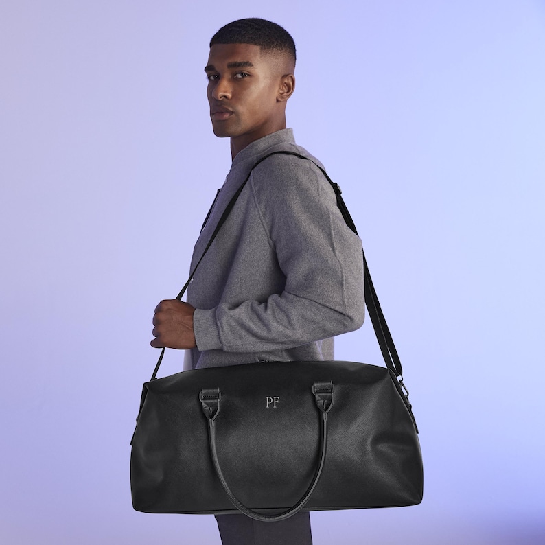 Puede incluir: Una bolsa de viaje de cuero negro con una correa para el hombro y dos asas. La bolsa tiene una cremallera y un monograma "PF" en la parte delantera.