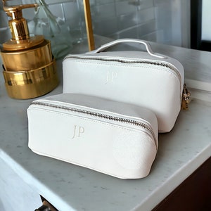 May include: Two off-white zippered cosmetic bags with gold zippers and the initials "JP" embossed on the front. The bags are on a white marble surface.