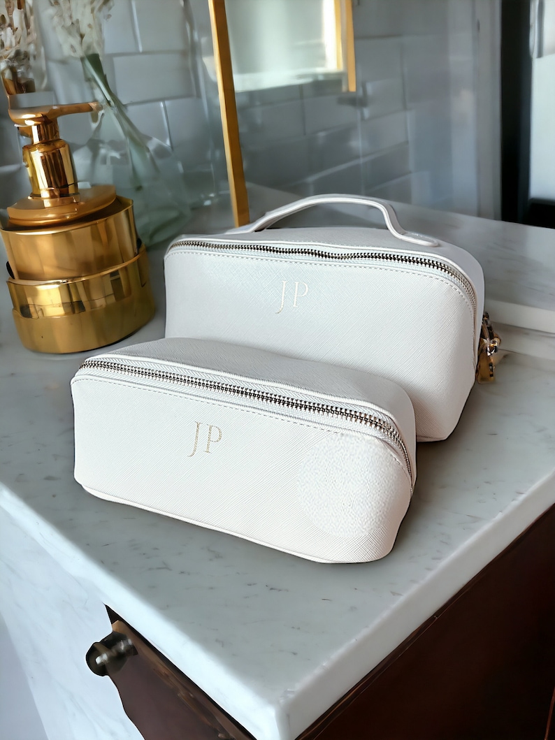 May include: Two off-white zippered cosmetic bags with gold zippers and the initials "JP" embossed on the front. The bags are on a white marble surface.