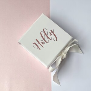 K&ouml;nnte beinhalten: Eine wei&szlig;e Geschenkbox mit einem wei&szlig;en Satinband, das zu einer Schleife gebunden ist. Die Box tr&auml;gt den Namen "Holly" in ros&eacute;goldenen Lettern.