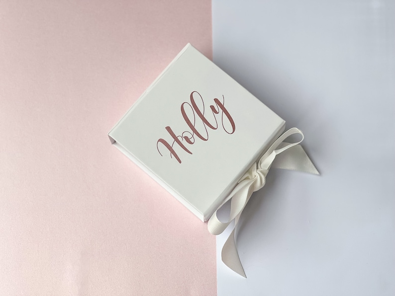 K&ouml;nnte beinhalten: Eine wei&szlig;e Geschenkbox mit einem wei&szlig;en Satinband, das zu einer Schleife gebunden ist. Die Box tr&auml;gt den Namen "Holly" in ros&eacute;goldenen Lettern.