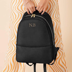 Può includere: Zaino in pelle nera con tiretti delle cerniere dorati e un monogramma dorato che recita "N.B."