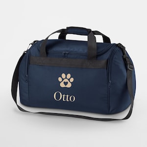 Peut inclure: Sac de sport bleu marine avec poignées et bandoulière noires. Le sac présente une empreinte de patte beige avec un cœur et le nom "Otto" imprimé en dessous. Le sac a un dessus zippé et des poches latérales.