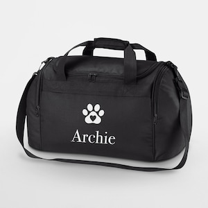 Peut inclure: Sac de sport noir avec une empreinte de patte blanche et un cœur, et le nom "Archie" en dessous. Le sac a une poignée supérieure, une bandoulière et des poches latérales. Idéal pour le sport ou les voyages.