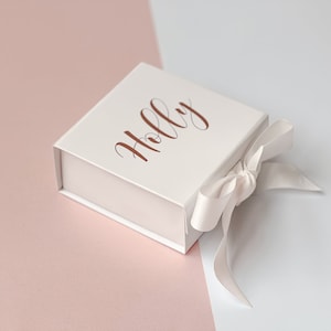 K&ouml;nnte beinhalten: Eine wei&szlig;e Geschenkbox mit einem ros&eacute;goldenen Band und dem Namen "Holly" auf der Oberseite.