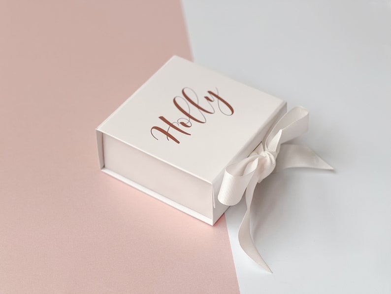 K&ouml;nnte beinhalten: Eine wei&szlig;e Geschenkbox mit einem ros&eacute;goldenen Band und dem Namen "Holly" auf der Oberseite.