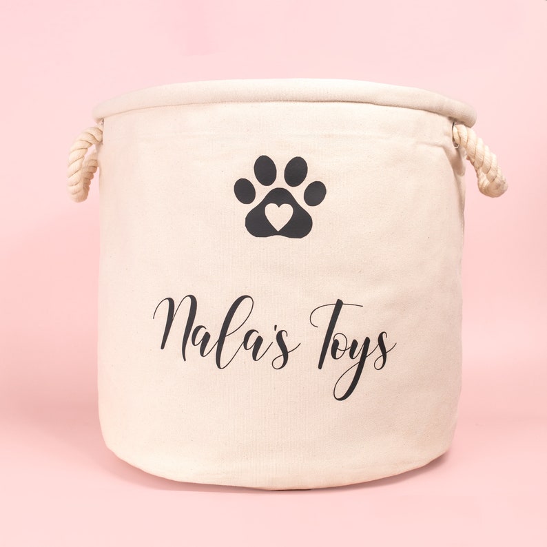 May include: A cylindrical off-white canvas storage basket featuring two thick twisted rope handles on the sides. The front displays a black graphic of a paw print with a heart-shaped center and the black cursive text Nala's Toys below it.