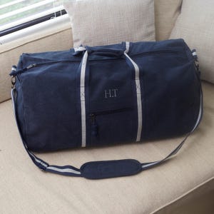 Puede incluir: Una bolsa de lona azul marino con rayas blancas en las asas y un bolsillo con cremallera en la parte delantera. La bolsa tiene una correa para el hombro y est&aacute; monogramada con las iniciales "H.T."