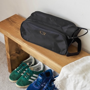 Puede incluir: Bolsa de zapatos negra con cierre de cremallera y asa. La bolsa tiene las iniciales "CP" bordadas en la parte delantera. La bolsa est&aacute; sentada en un banco de madera.
