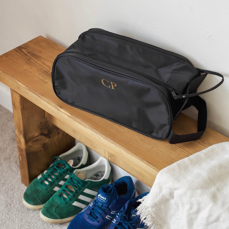 Puede incluir: Bolsa de zapatos negra con cierre de cremallera y asa. La bolsa tiene las iniciales "CP" bordadas en la parte delantera. La bolsa est&aacute; sentada en un banco de madera.