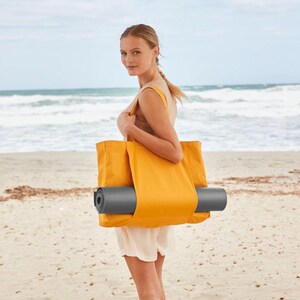 May include: A woman carrying a yellow tote bag with a rolled-up grey yoga mat inside. The bag has a wide, flat base and two handles. The woman is standing on a sandy beach with the sea in the background.