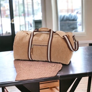 Puede incluir: Una bolsa de lona de color beige con correas a rayas marrones y blancas. La bolsa tiene un bolsillo con cremallera en la parte delantera y las iniciales "HC" bordadas en la parte delantera.