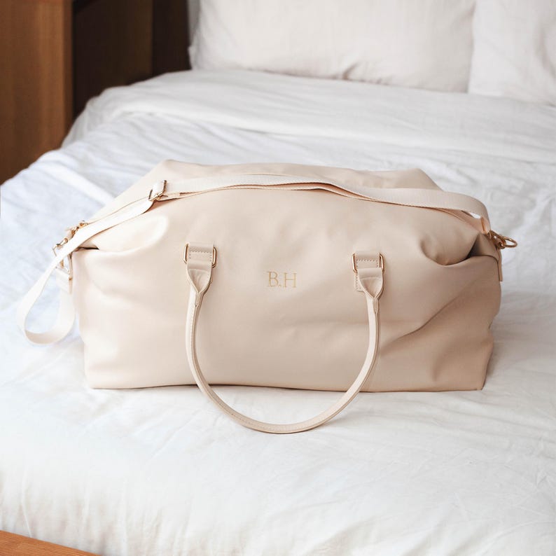 Puede incluir: Una bolsa de viaje de cuero color crema con herrajes dorados y un monograma "B.H." en la parte delantera.