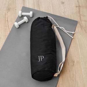 May include: Black yoga mat bag with a white drawstring and a white strap. The bag has the initials "JP" embroidered on the front.