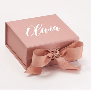 K&ouml;nnte beinhalten: Eine Geschenkbox in Blush-Rosa mit einem wei&szlig;en Satinband und dem Namen "Olivia" auf der Oberseite.