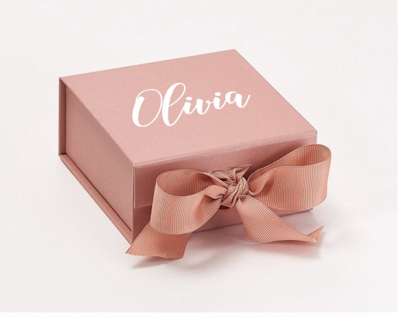 K&ouml;nnte beinhalten: Eine Geschenkbox in Blush-Rosa mit einem wei&szlig;en Satinband und dem Namen "Olivia" auf der Oberseite.