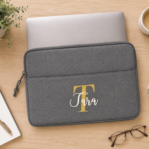 Könnte beinhalten: Eine anthrazitfarbene Laptoptasche aus Stoff mit einem großen goldenen Buchstaben T und dem Namen Tara in weißer Schreibschrift auf der Vordertasche. Die Tasche hat einen schwarzen Reißverschluss und eine rechteckige Form mit abgerundeten Ecken.