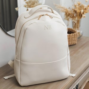 May include: Cream-colored backpack with gold-toned zippers and handle. The backpack has a rounded shape with a top handle and adjustable straps. The initials "NB" are subtly embossed on the front. The bag is sitting on a wooden surface.