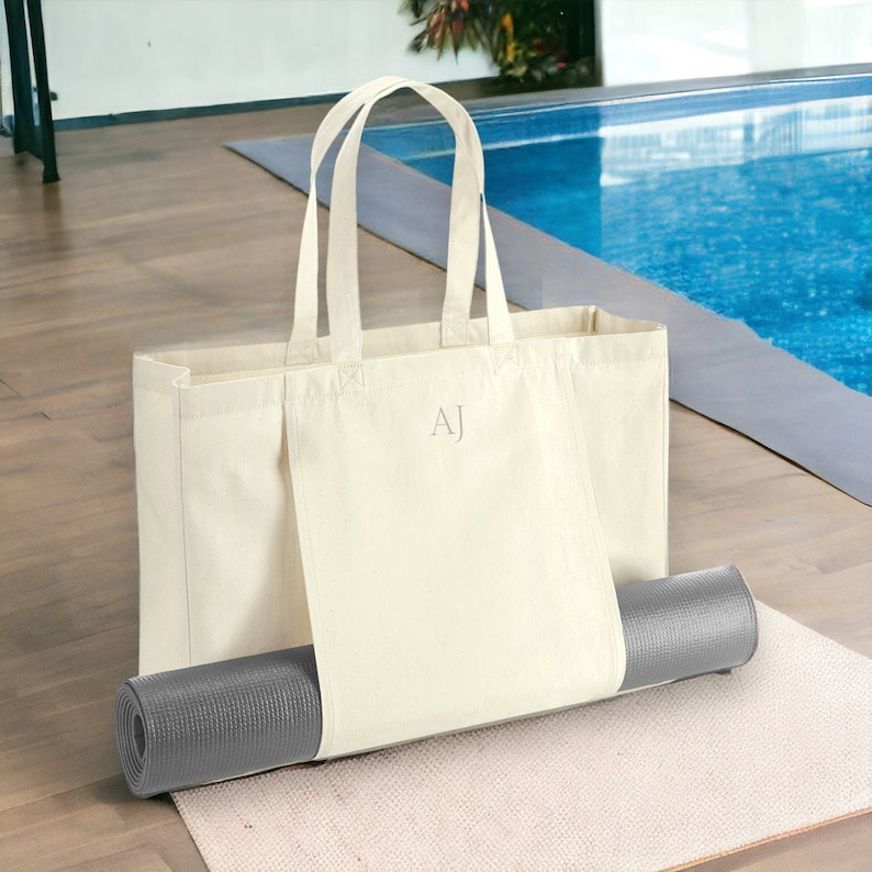 May include: A white canvas tote bag with the initials "AJ" embroidered on the front. The bag has a gray yoga mat tucked into the side pocket.