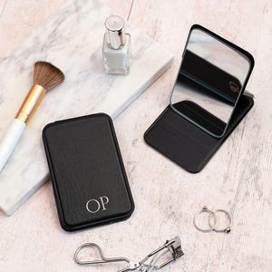 May include: A black, rectangular compact mirror with a silver initial monogram, open and closed, with a makeup brush, nail polish, rings, and eyelash curler on a light wood surface.
