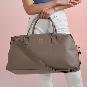 Puede incluir: Una bolsa de viaje de lona gris con una correa para el hombro y herrajes dorados. La bolsa tiene las iniciales "HG" grabadas en la parte delantera.