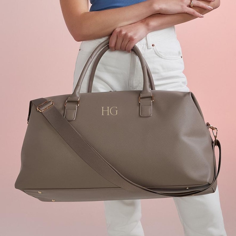 Puede incluir: Una bolsa de viaje de lona gris con una correa para el hombro y herrajes dorados. La bolsa tiene las iniciales "HG" grabadas en la parte delantera.