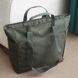Op de afbeelding: Een donkergroene canvas tote bag met twee bovenste handvatten en een ritsvak. De tas heeft de initialen "KC" geborduurd op de voorkant.