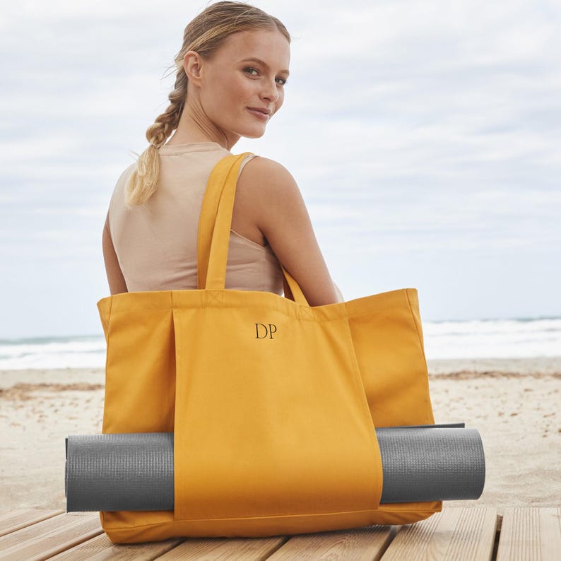 May include: A yellow canvas tote bag with a black monogram "DP" on the front. The bag has a pocket on the side that holds a gray yoga mat.