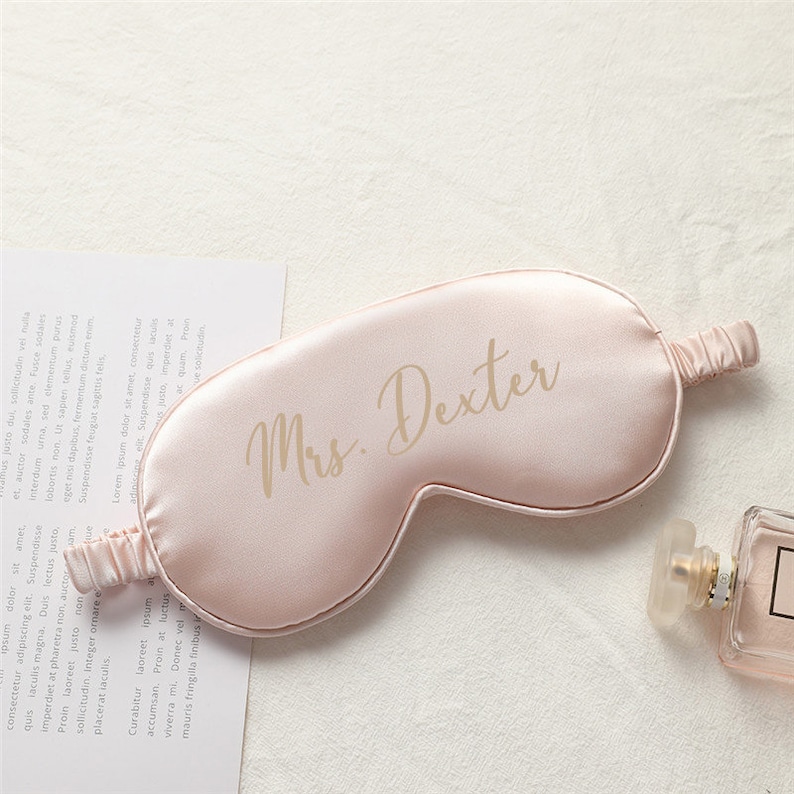 以下が含まれることがあります： ゴールドの刺繍で「Mrs. Dexter」と刺繍された、薄いピンクのサテン製のアイマスクです。